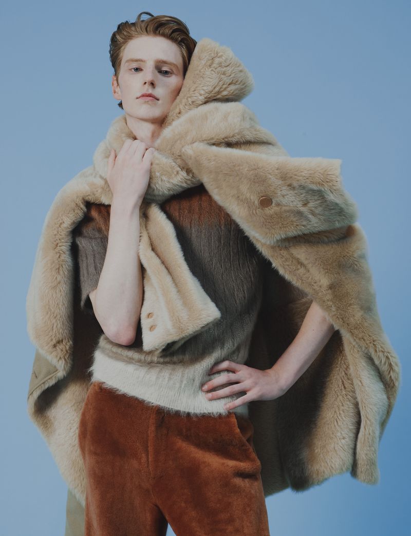 Fashionable ginger-haired person wearing a plush beige shawl and rust-colored pants in a studio shot against a light blue background.