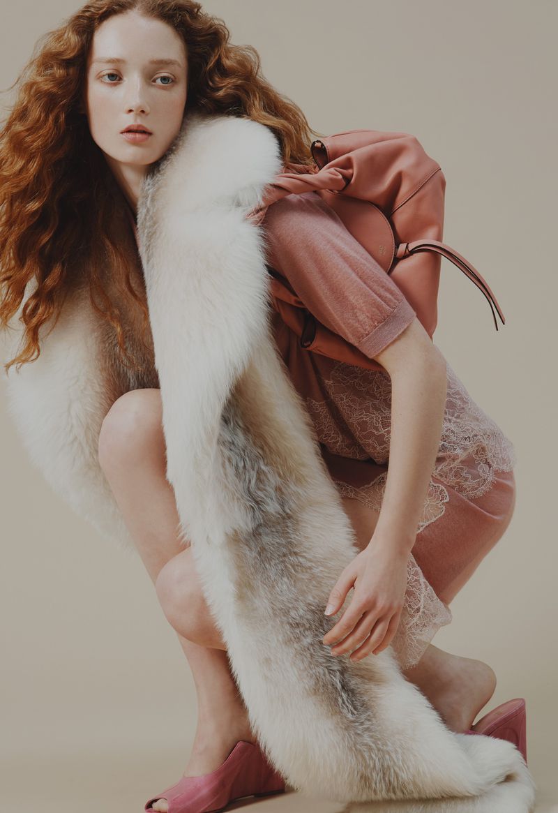 Fashionable woman with long red hair poses indoors wearing a short pink dress, pink shoes, and a long white faux fur stole.