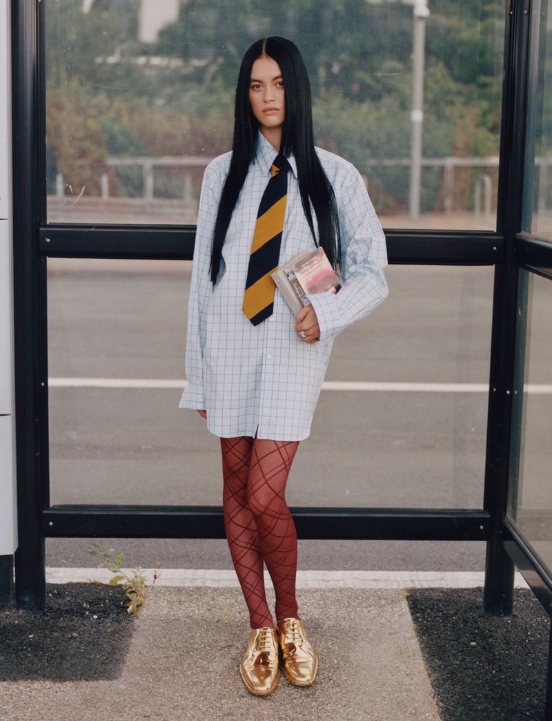A young adult woman in patterned shirt and tie, stands at a bus stop with glossy shoes, holding books in an outdoor setting.