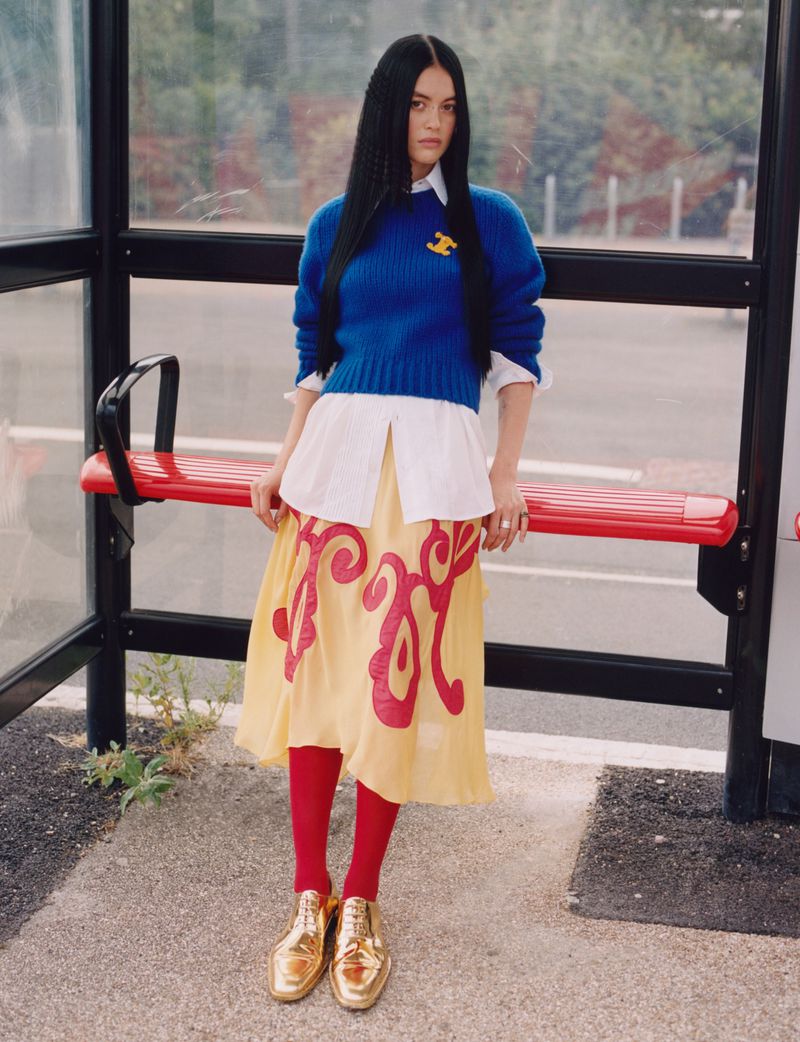 An adult female stands outside a bus stop wearing a blue sweater, white button-down shirt, and a yellow skirt with red tights and gold shoes.