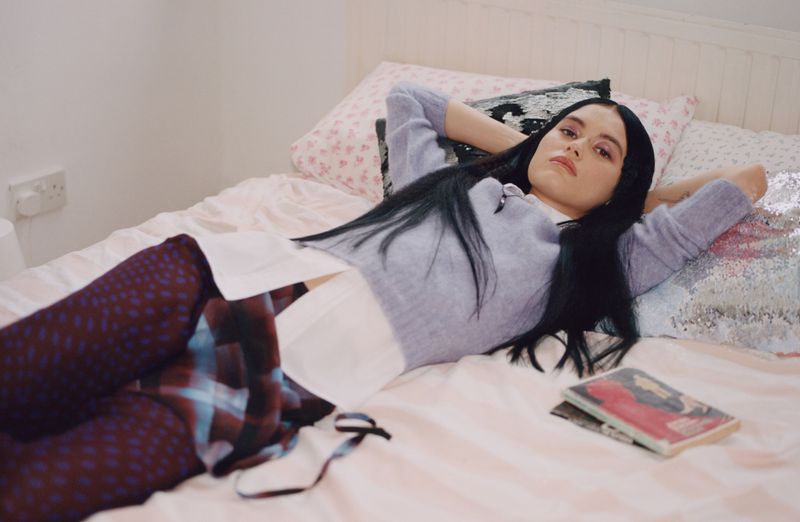 Young woman with long dark hair lounges on a pink bed in a lavender sweater with books and patterned leggings in an indoor setting.