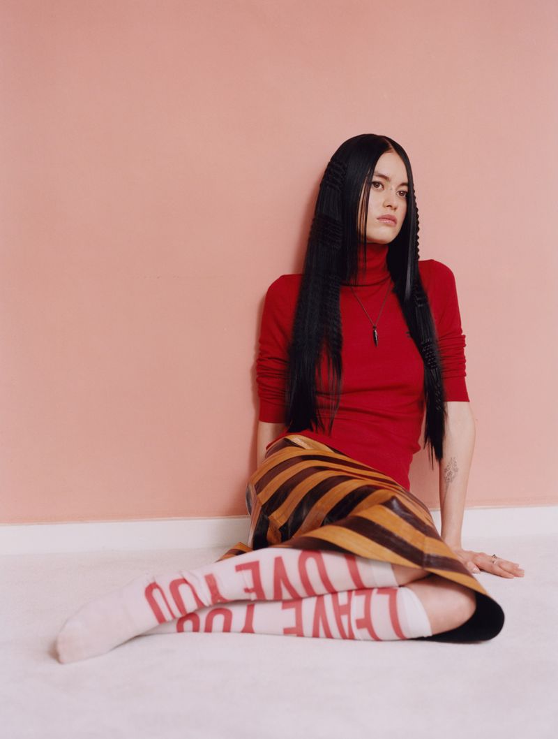 A woman with long black hair sits against a pink wall wearing a red turtleneck, striped skirt, and white socks with red text.