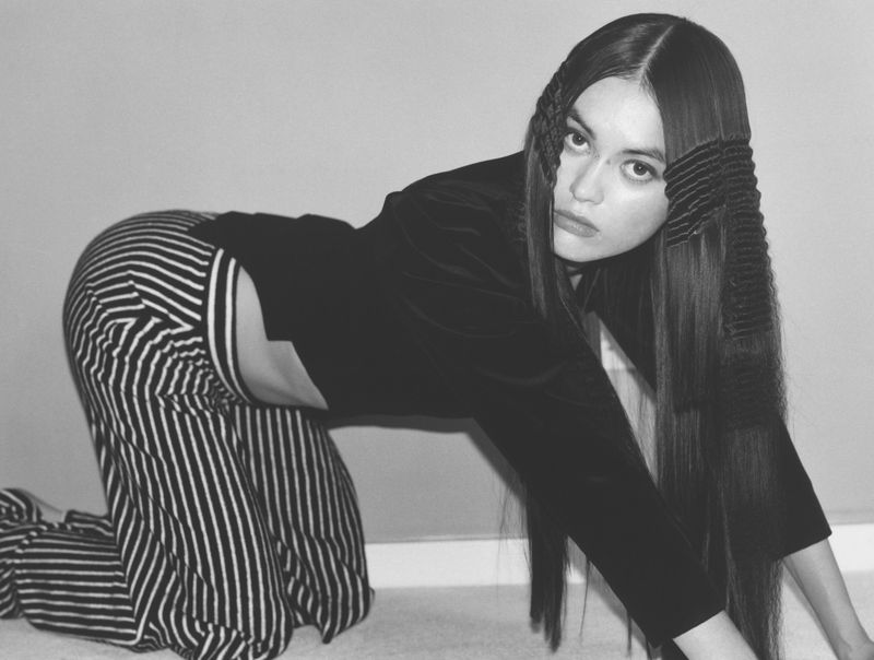 Black and white portrait of a woman with long dark hair kneeling in striped pants and a black top in front of a wall.