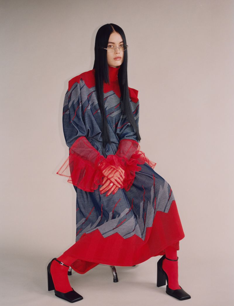 Woman with long black hair poses seated in a blue and red fashion dress, wearing black heeled shoes, against a light background in a studio.