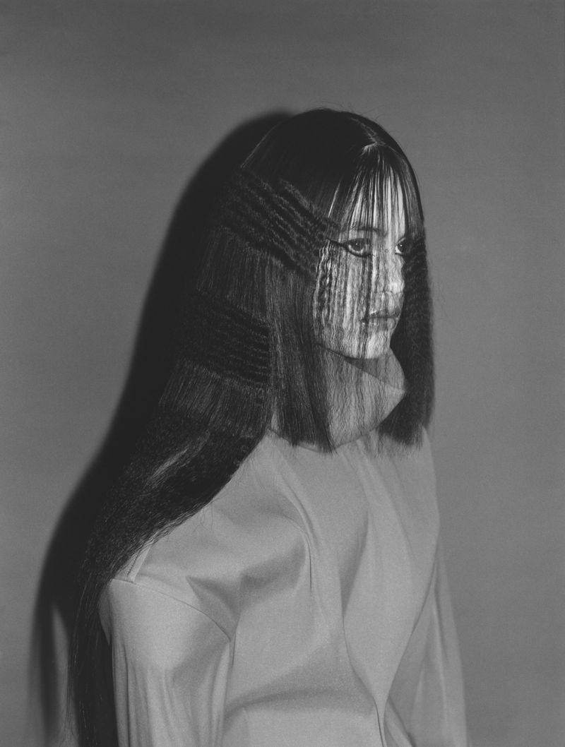 Monochrome portrait of an adult woman with long dark hair, styled with crimped texture and wearing a light-colored blouse against a neutral background.