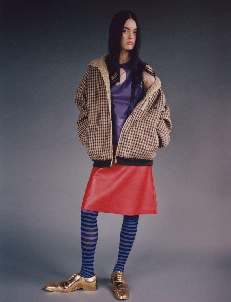 A woman in a plaid jacket, purple top, red skirt, and blue striped tights poses in a studio with gold shoes in a fashion shot.