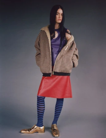 A woman in a plaid jacket, purple top, red skirt, and blue striped tights poses in a studio with gold shoes in a fashion shot.