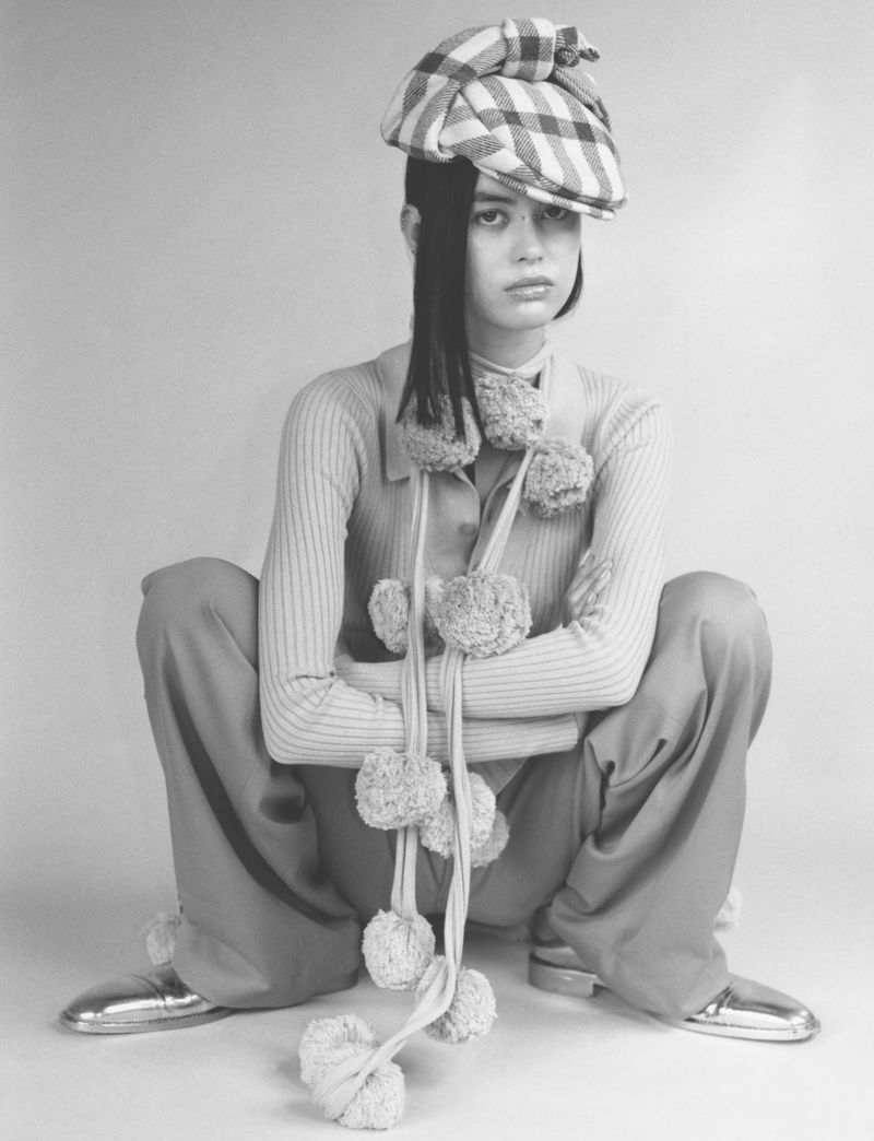 Fashionable young adult in a checked cap and pom-pom scarf poses in studio, wearing coordinated pale top, trousers, and metallic shoes.