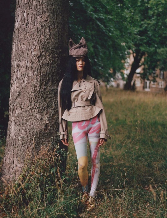 Model leaning against a large tree trunk wearing a beige cropped trench coat, patterned leggings, and a distinctive crocheted hat.