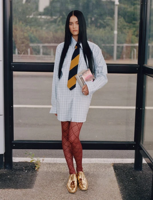 Model with long dark hair wearing an oversized plaid shirt, striped tie, and red patterned tights at a bus shelter.