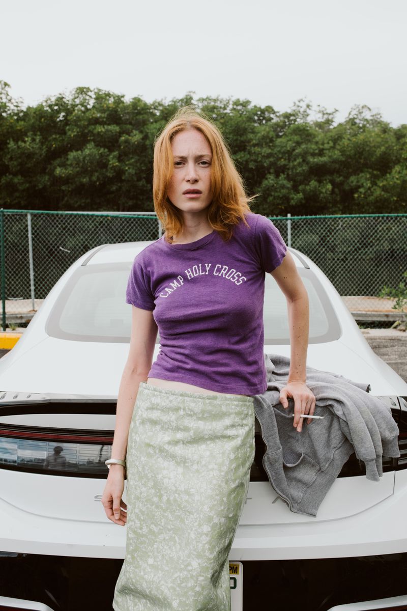 Woman with auburn hair poses against a white car, wearing a purple t-shirt and green skirt, holding a cigarette in a parking lot.