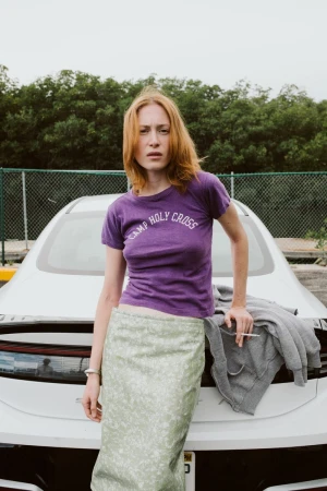 Woman with auburn hair poses against a white car, wearing a purple t-shirt and green skirt, holding a cigarette in a parking lot.