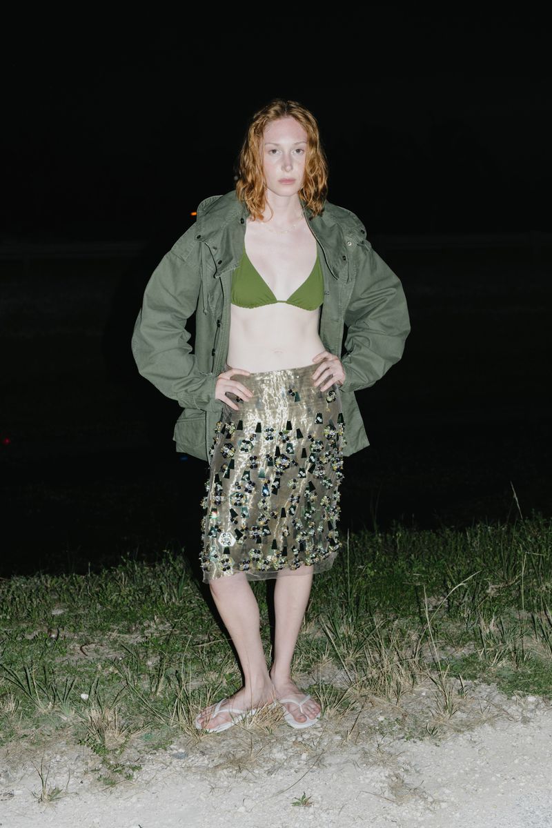 Woman in bikini top, khaki jacket, and sequin skirt standing in a dark outdoor location with grass and partial water view.