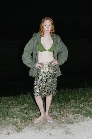 Woman in bikini top, khaki jacket, and sequin skirt standing in a dark outdoor location with grass and partial water view.