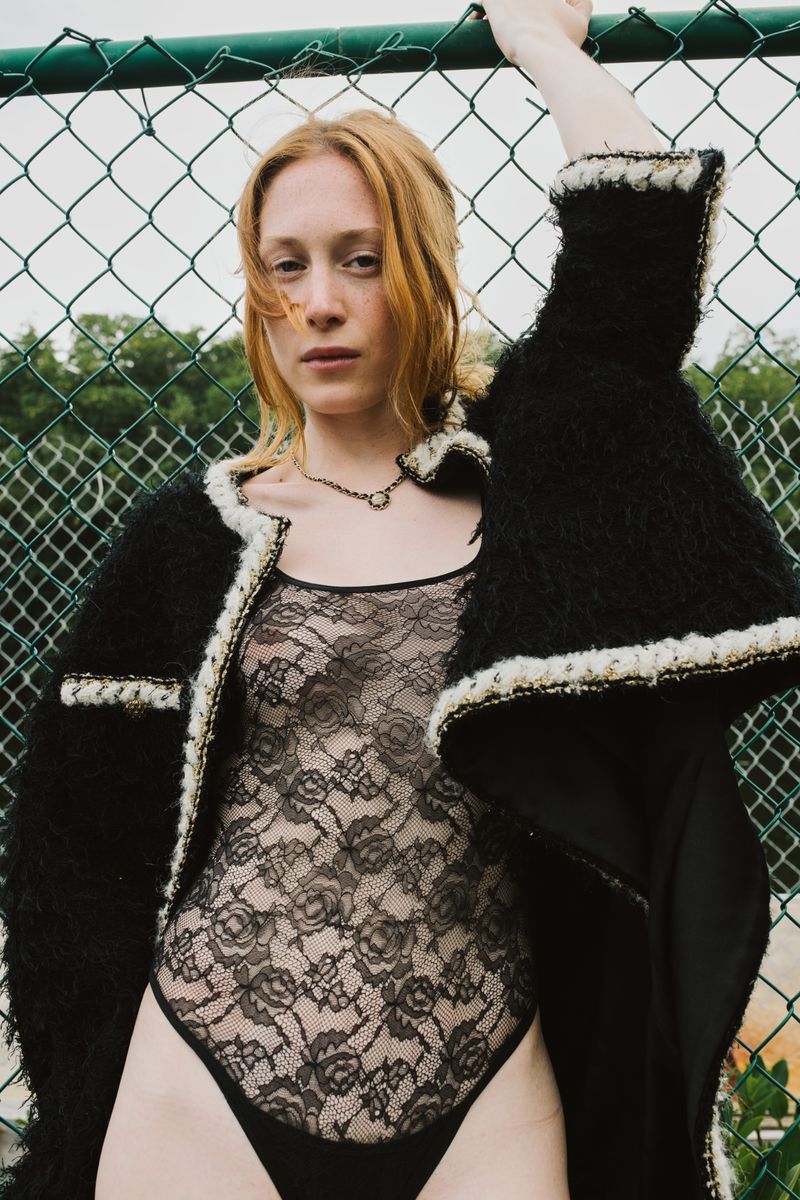 A fashionable, red-haired adult woman in a black lace bodysuit and black jacket poses against a chain-link fence in an outdoor setting.