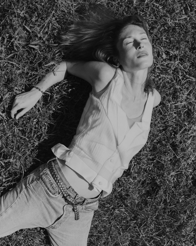 A woman lies on her back in the grass with her eyes closed, wearing jeans and a vest in this high angle black and white photo.