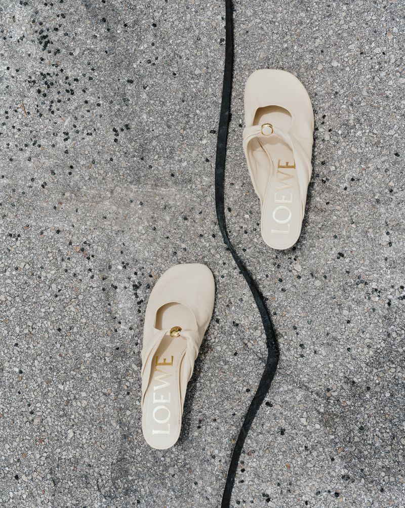 Overhead shot of two cream-colored Loewe ballet flats with a black leather strap on a speckled grey concrete surface. Fashion product shot.