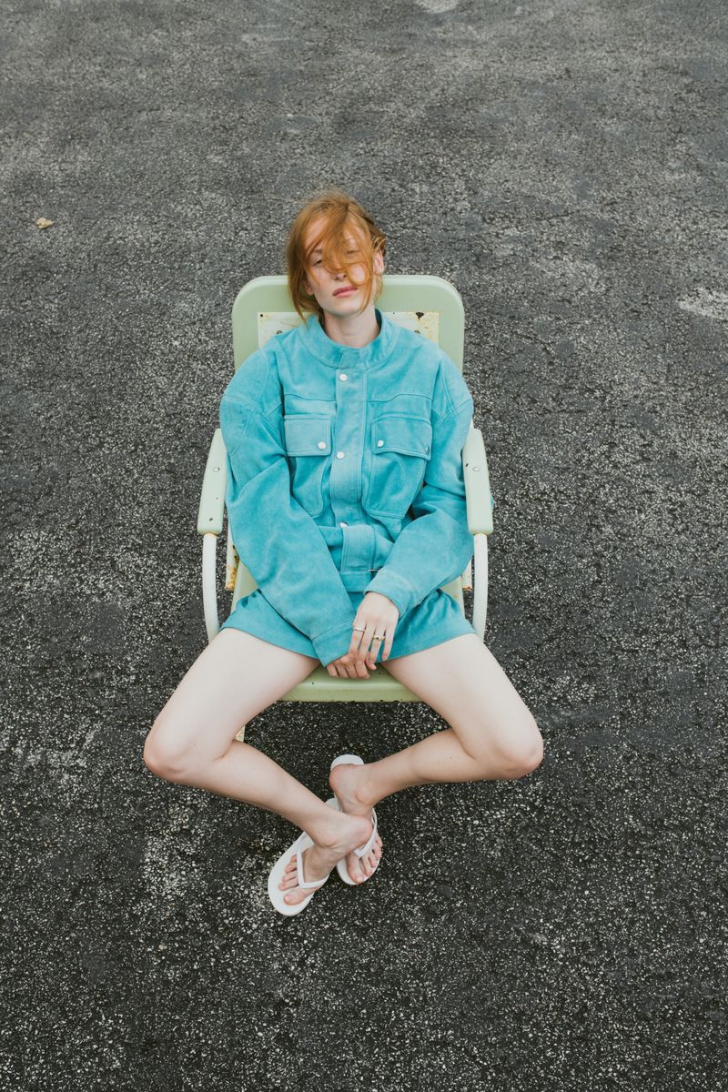 Red-haired adult woman in a turquoise shirt and shorts sits cross-legged in a green chair on an asphalt surface in an overhead shot.