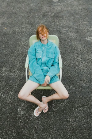 Red-haired adult woman in a turquoise shirt and shorts sits cross-legged in a green chair on an asphalt surface in an overhead shot.
