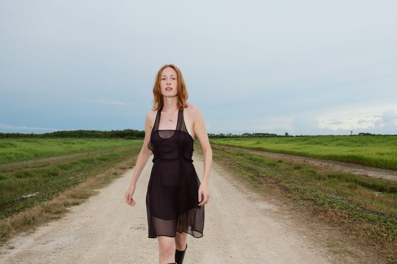 A woman in a black dress walks along a dirt road, backed by a green field under a cloudy sky, captured in a medium shot.