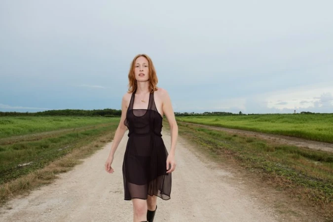 A woman in a black dress walks along a dirt road, backed by a green field under a cloudy sky, captured in a medium shot.