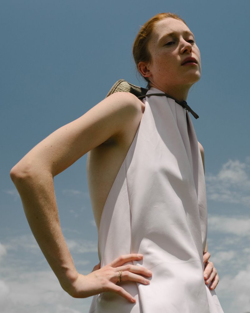 Low-angle shot of model Hunter Pifer with red hair wearing a pale halter top with hands on hips against an open blue sky.