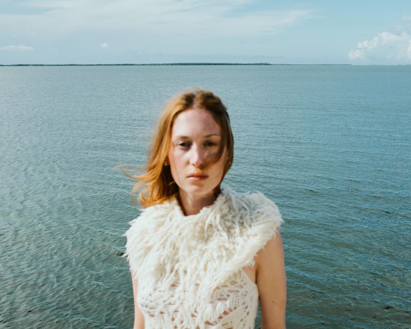 Woman poses in a knit top in front of a grey ocean and pale blue sky, creating a serene, natural fashion portrait.