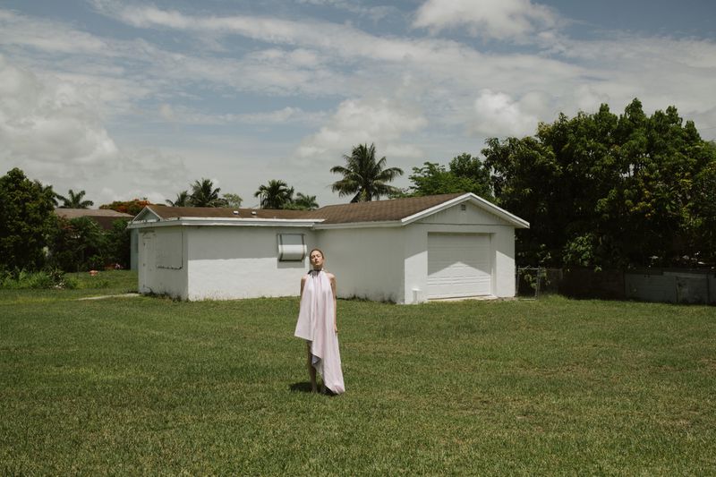 A woman stands in a grassy yard in a long, flowing white dress in front of a white single-story house on a partly cloudy day.
