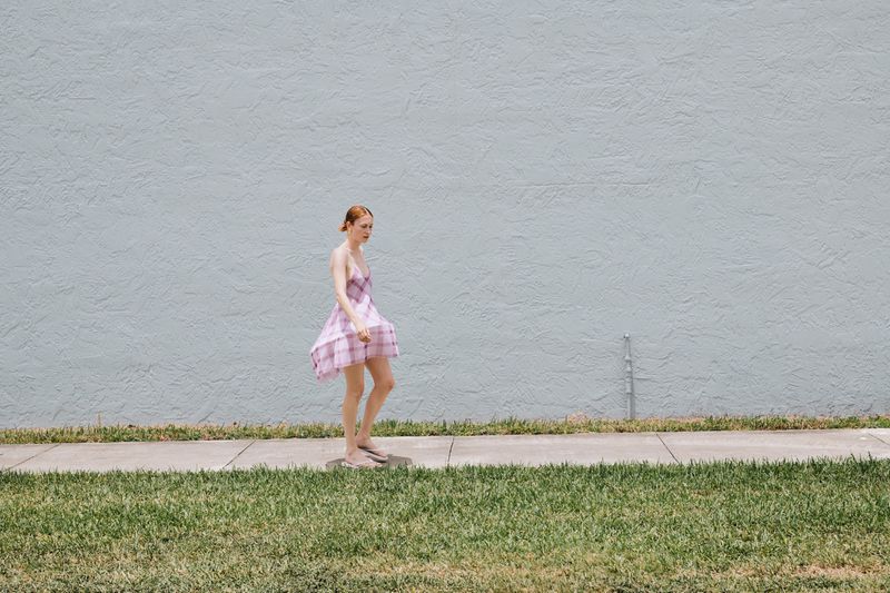 Adult female in dress walks beside concrete path and grass in a sunlit outdoor setting near textured light blue wall.