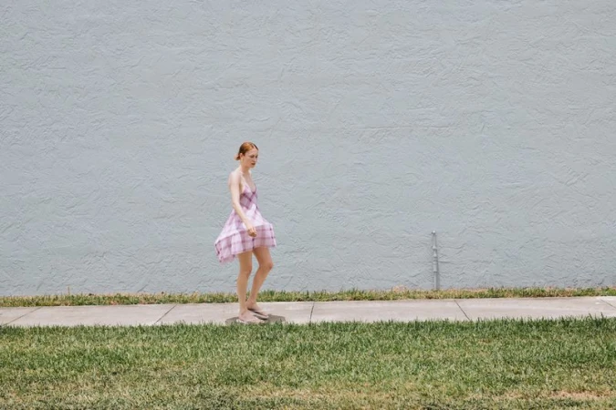 Adult female in dress walks beside concrete path and grass in a sunlit outdoor setting near textured light blue wall.