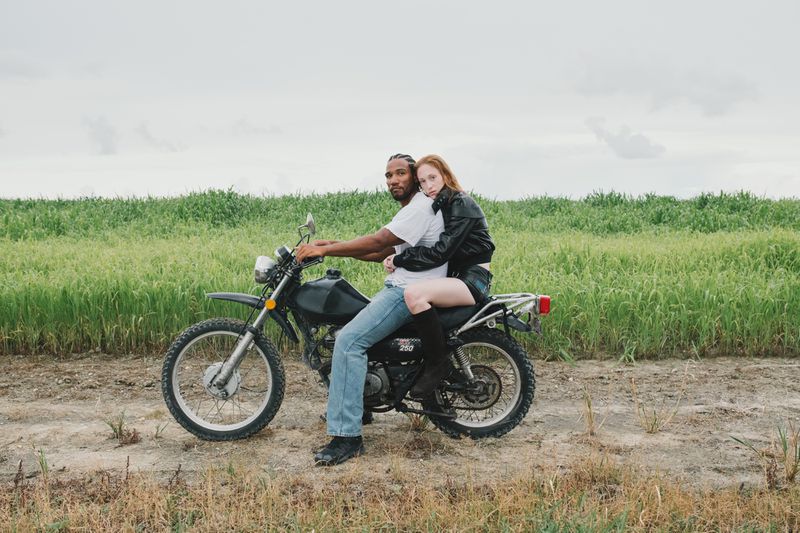 Man and woman on a motorcycle in a rural field. The couple is sitting on the bike with a green field and sky in the background.