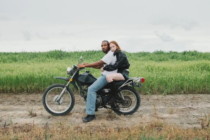 Man and woman on a motorcycle in a rural field. The couple is sitting on the bike with a green field and sky in the background.