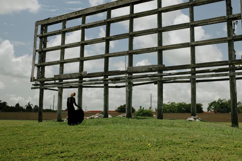 Adult female in a black dress poses in a grassy field near an abandoned billboard structure, under a partly cloudy sky.