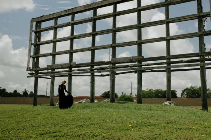 Adult female in a black dress poses in a grassy field near an abandoned billboard structure, under a partly cloudy sky.