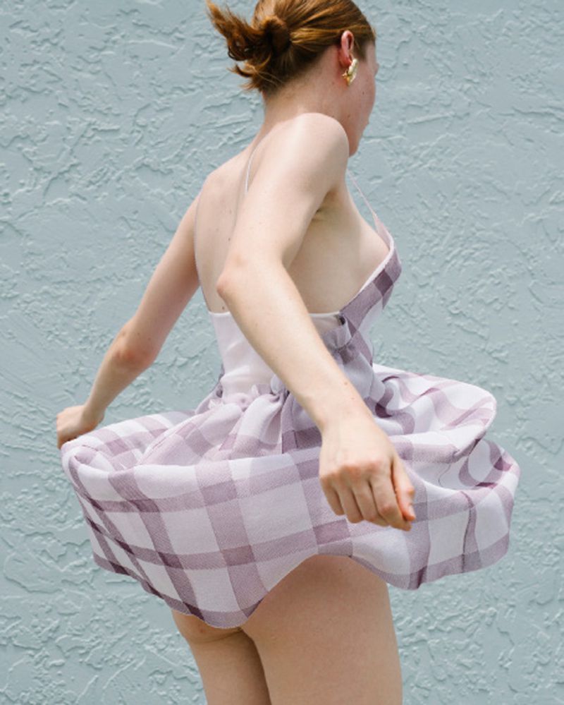 Woman in a sundress twirls in front of a blue wall. The dress is plaid. She wears gold earrings. The image is taken in daylight.