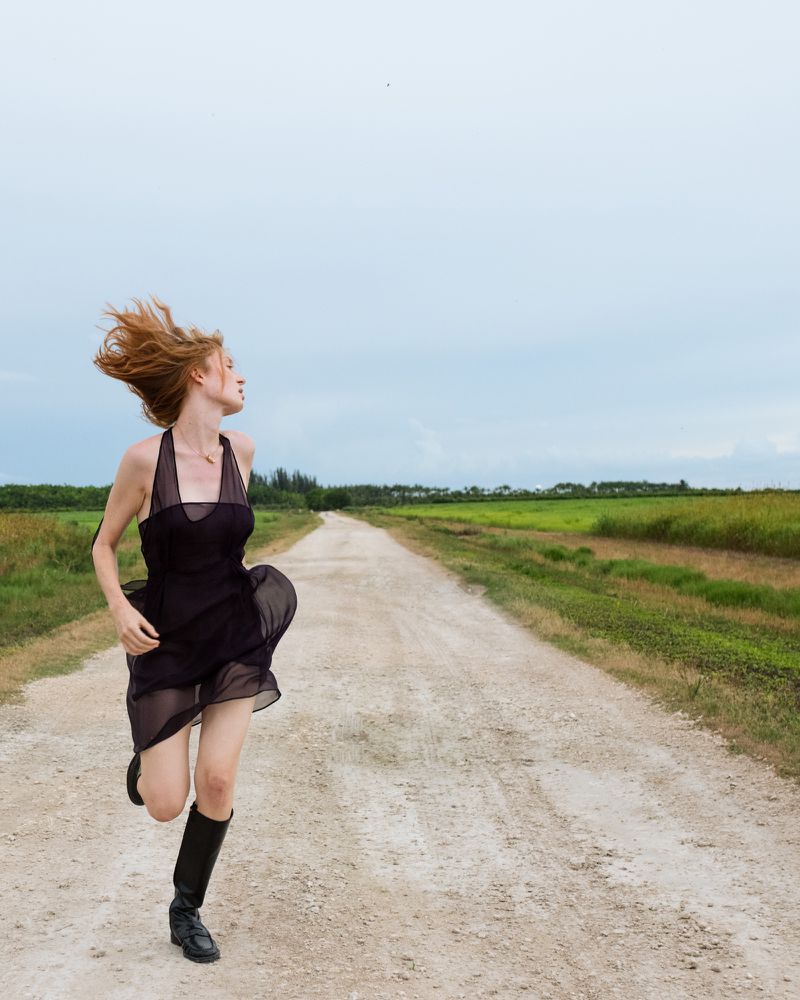 Red-haired woman in a black dress and boots runs along a dirt road in a rural landscape under an overcast sky, hair blowing in the wind.