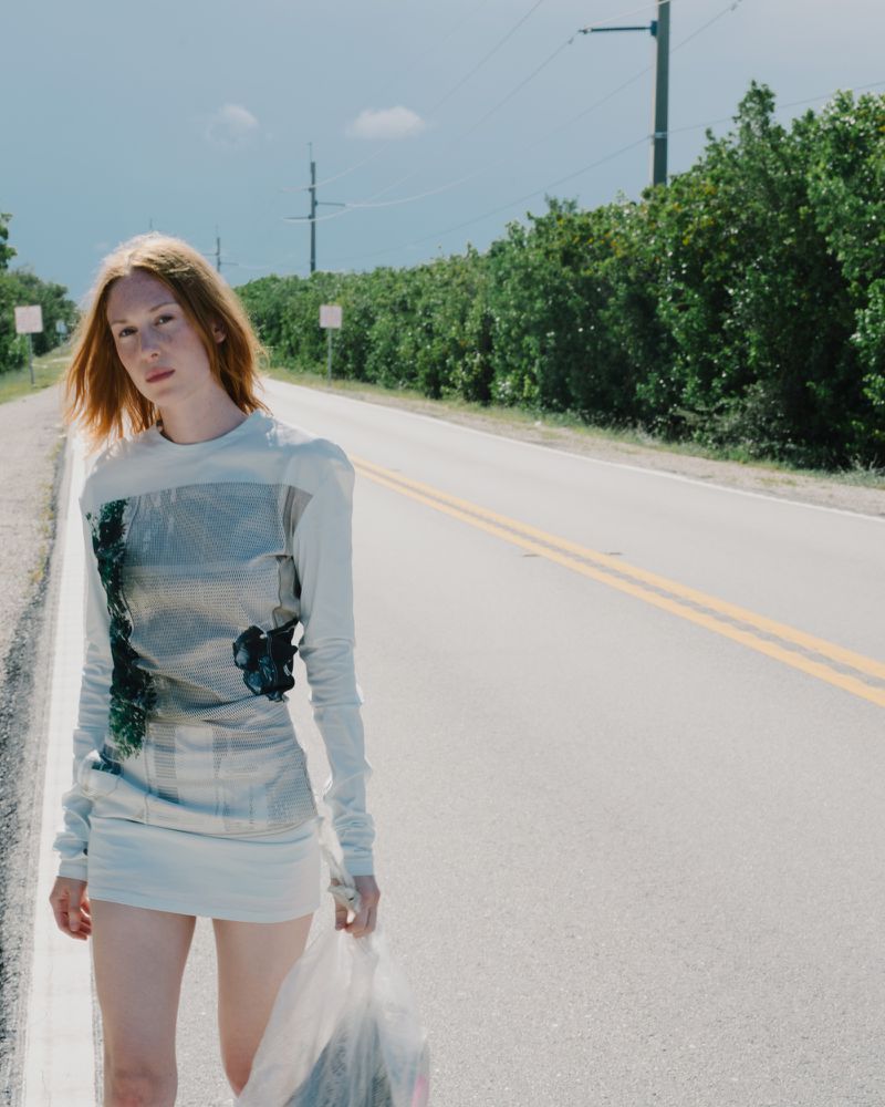 A redhead fashion model in a dress walks on an empty road with green trees and a cloudy blue sky on the horizon.