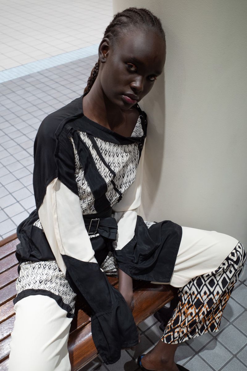 A striking fashion portrait of a dark-skinned woman with braided hair in a stylish black and white patterned outfit, leaning on a pillar.
