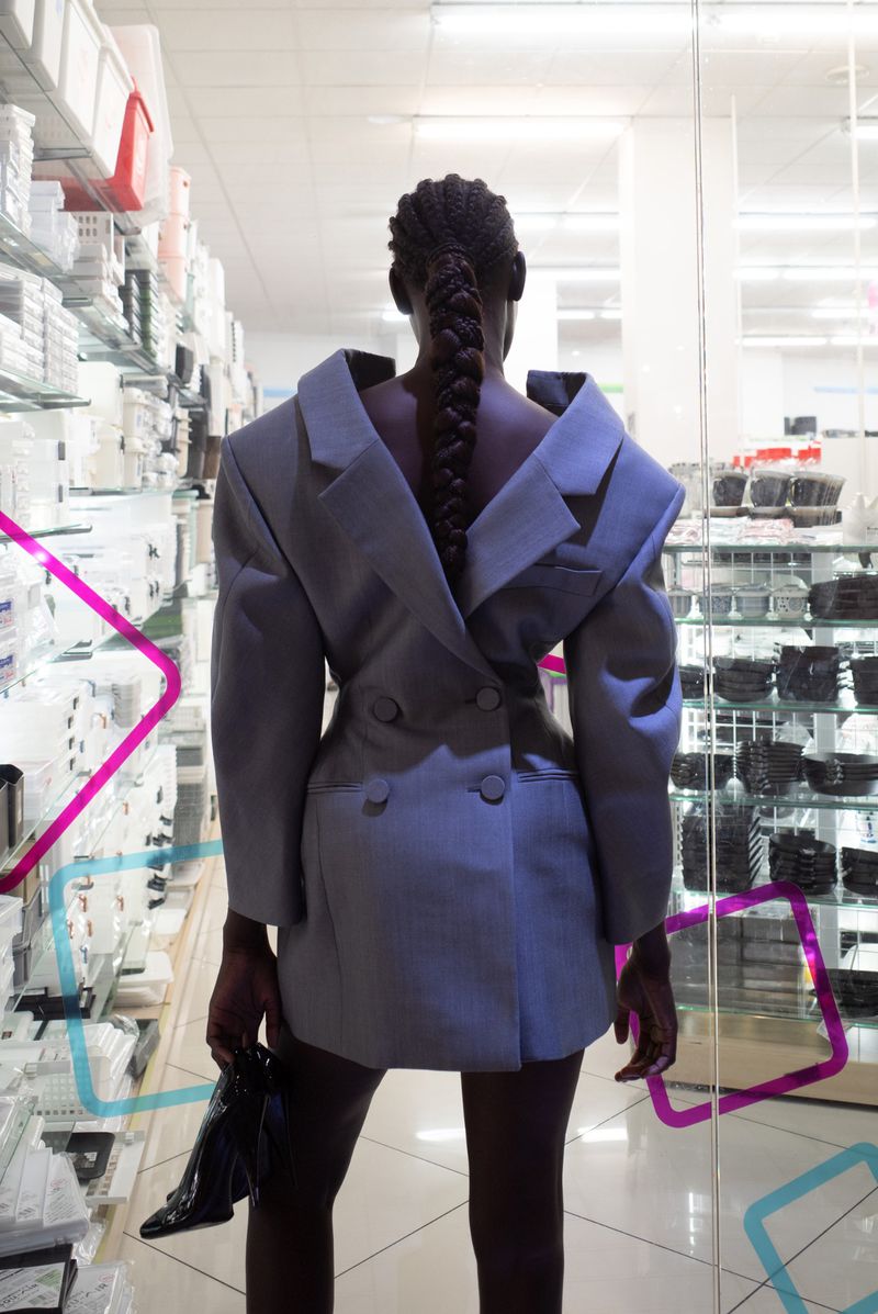 Woman in oversized grey blazer dress stands in front of shelves in an indoor setting, facing away from the camera, holding black shoes.