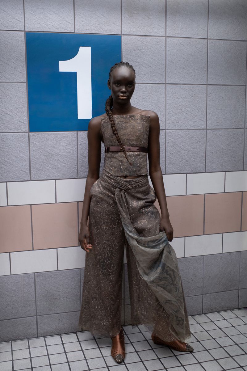 Elegant Black model poses in textured bronze outfit near subway number sign and tiled wall, showcasing high-fashion style.