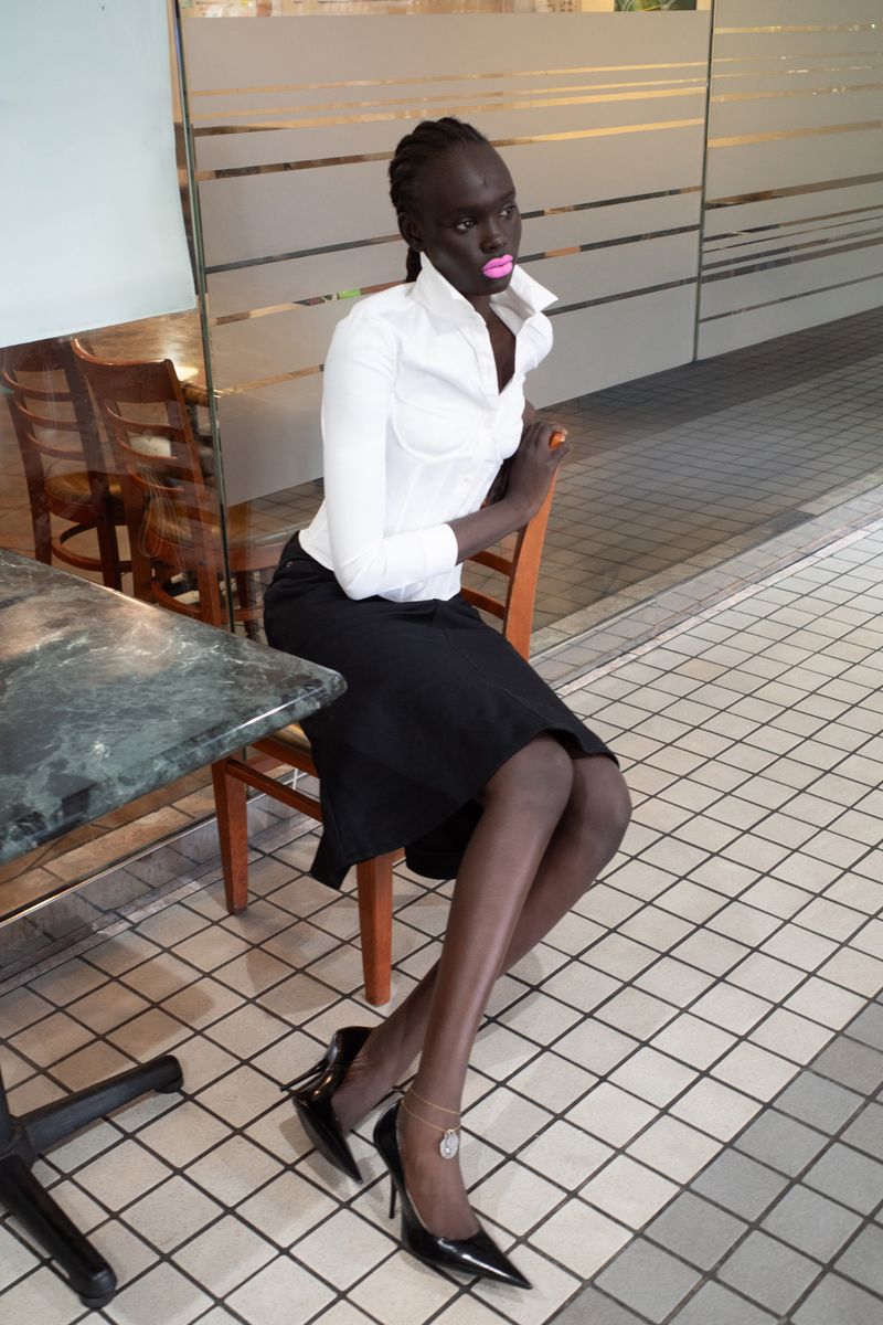 Elegant dark-skinned woman in white shirt and black skirt sits on a chair indoors with a marble table and tile floor.