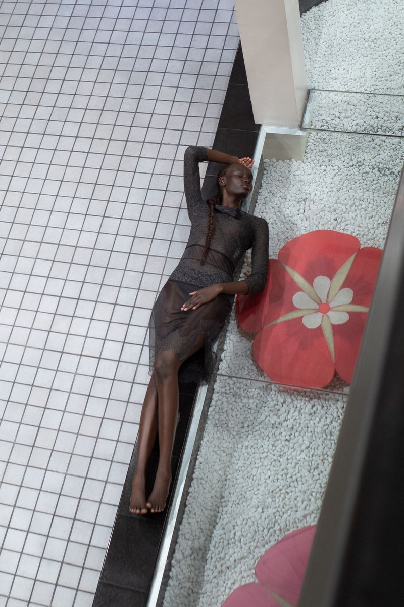 Dark-skinned woman in sheer dress lies on a textured surface near flower pillows. An overhead shot shows the model in a fashion pose.
