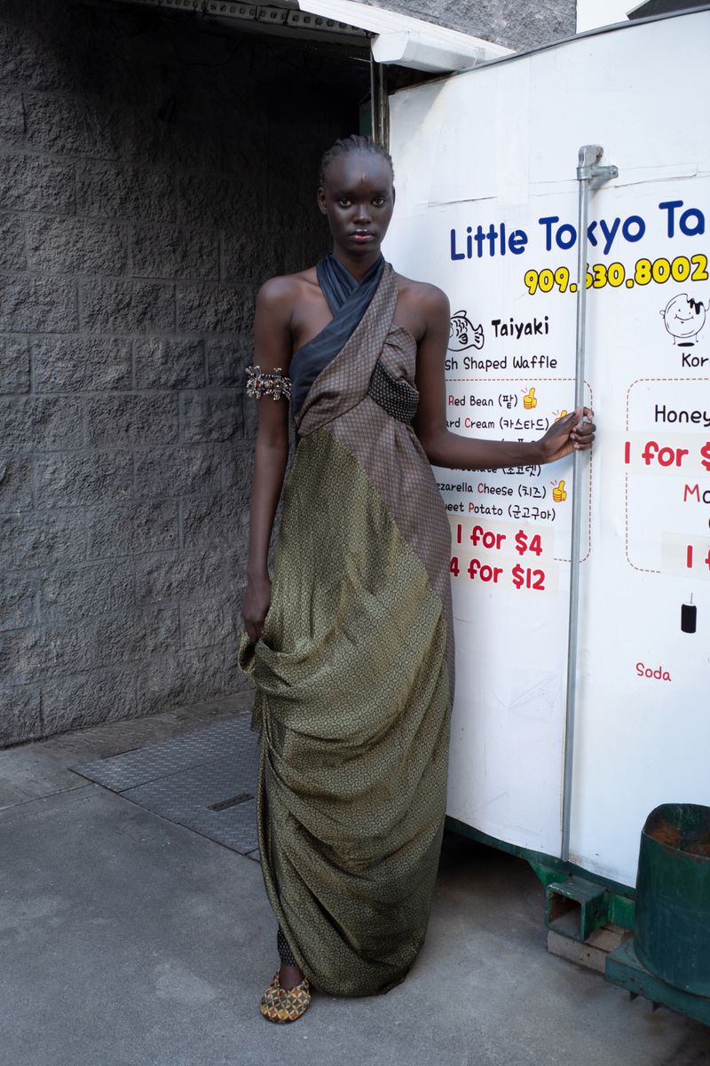 A statuesque woman in an earth-toned gown poses next to a food cart featuring a 'Little Tokyo Taiyaki' menu in an urban outdoor setting.