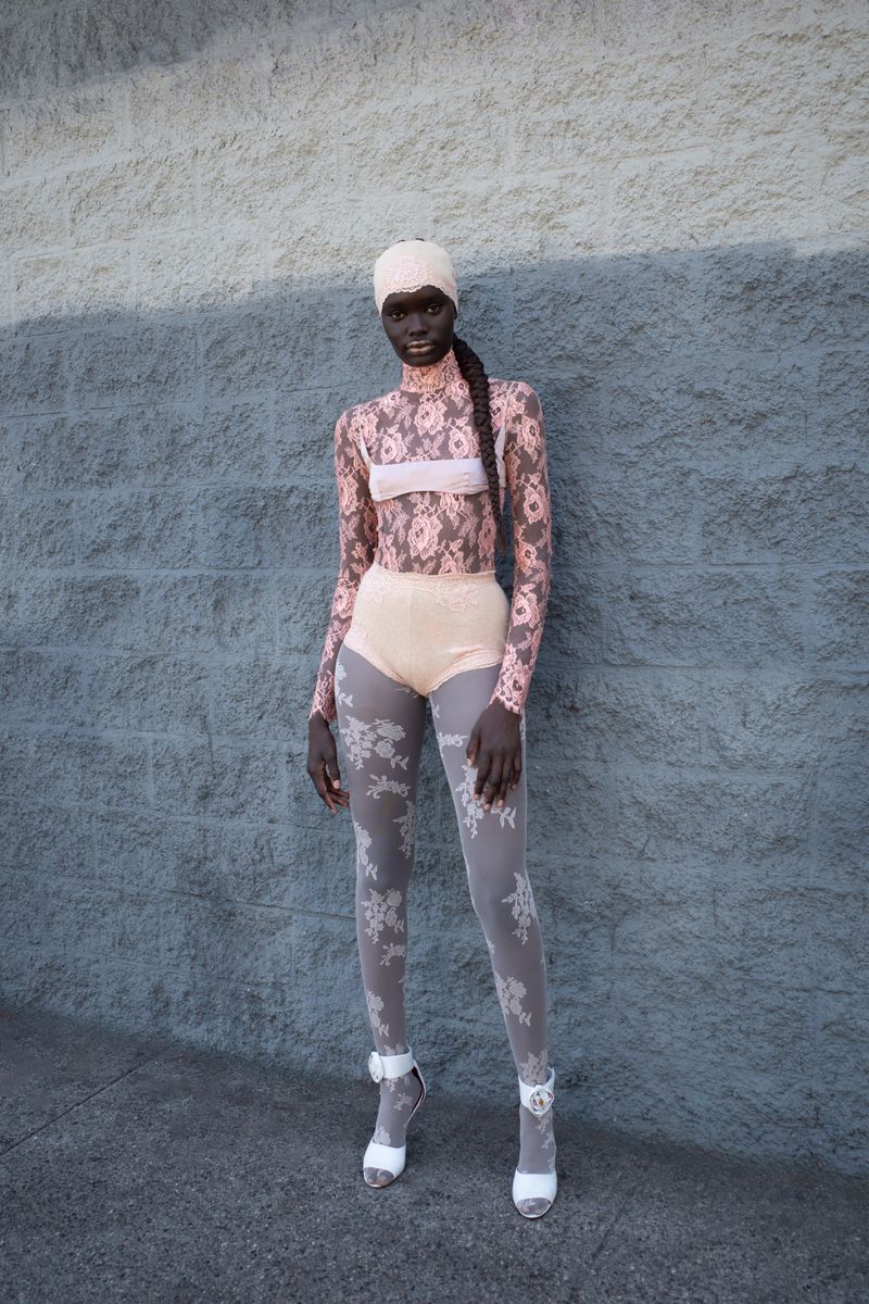 A slender woman poses in a high-fashion, rose-patterned bodysuit with matching beanie in front of a pale-blue cinderblock wall.