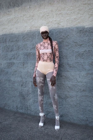 A slender woman poses in a high-fashion, rose-patterned bodysuit with matching beanie in front of a pale-blue cinderblock wall.