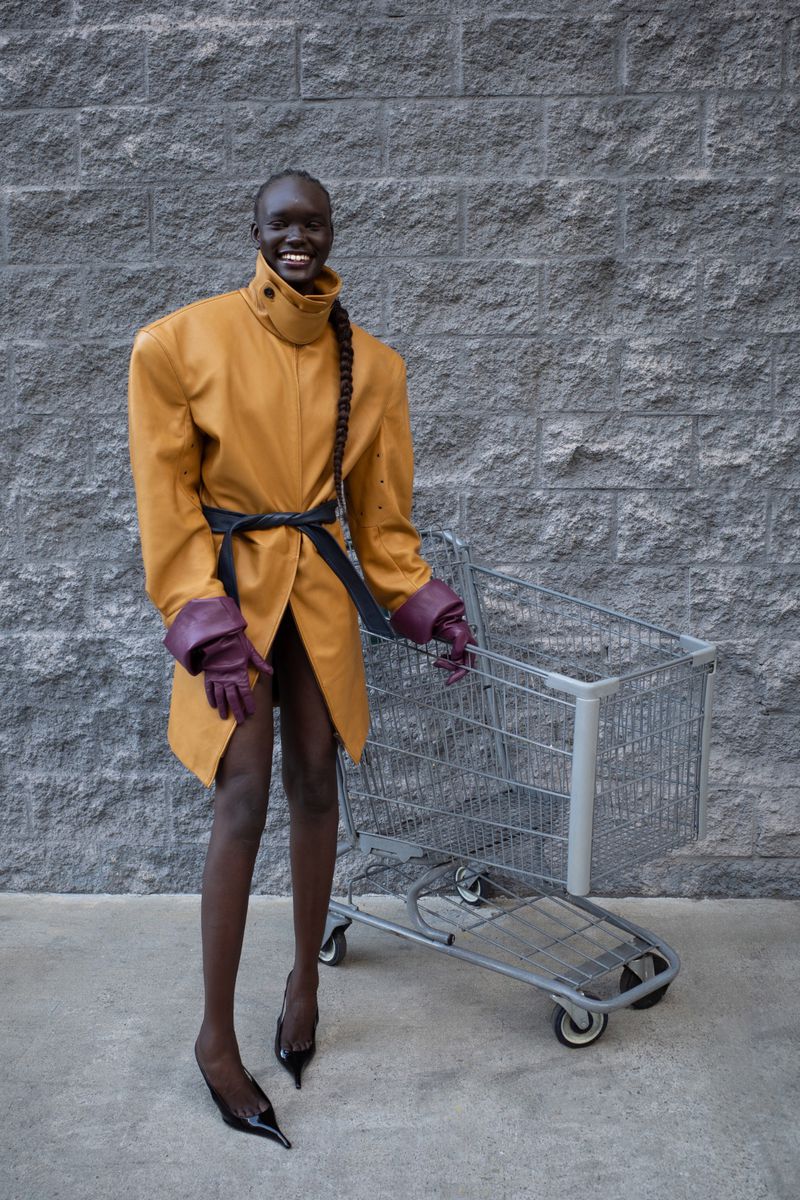 A slender adult female poses in a tan coat and heels, pushing a shopping cart against a grey brick wall, smiling slightly at the camera.