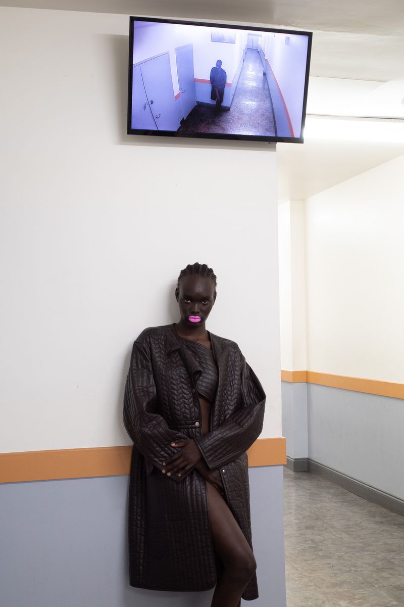 An adult female model with dark skin and braided hair posing in a black leather coat in a white corridor with a security monitor.