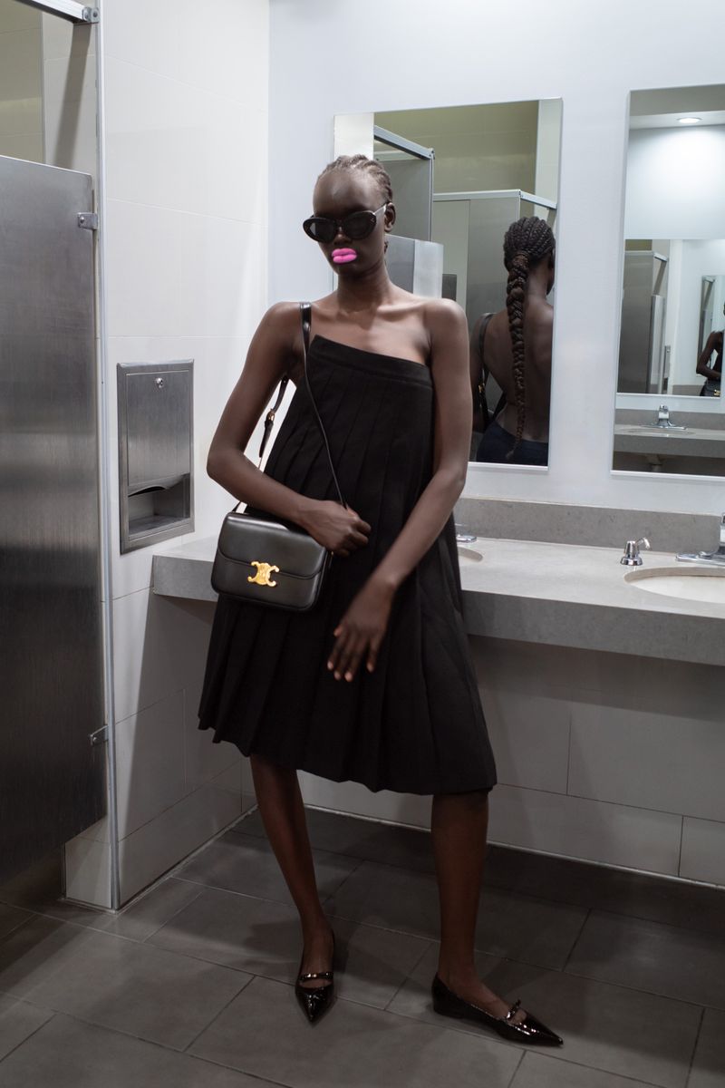 Woman in a black dress with sunglasses and a handbag stands in a bathroom. Braided hair is visible in a mirror behind her.