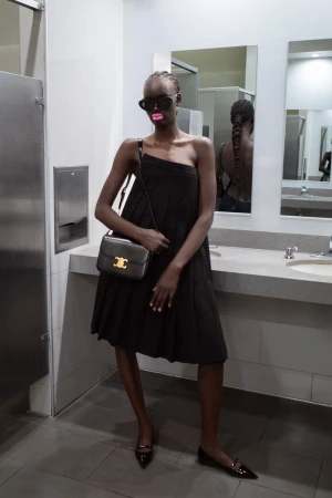Woman in a black dress with sunglasses and a handbag stands in a bathroom. Braided hair is visible in a mirror behind her.