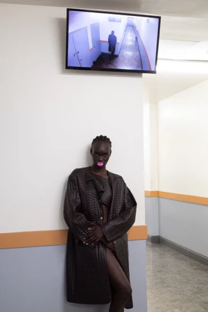 Black model with bright pink lipstick wearing a textured dark brown coat leaning against a wall beneath a security monitor screen.